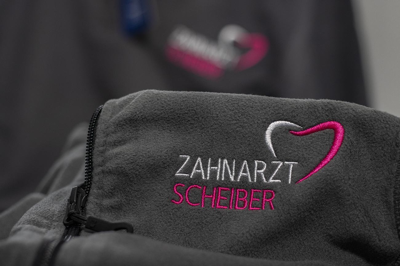 Dieselbe graue Fleecejacke mit gesticktem Praxislogo Zahnarzt Scheiber in Wei&szlig; und Magenta